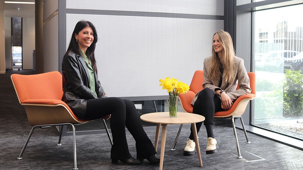 Two women talking at a small coffee table with vase of yellow flowers on top.