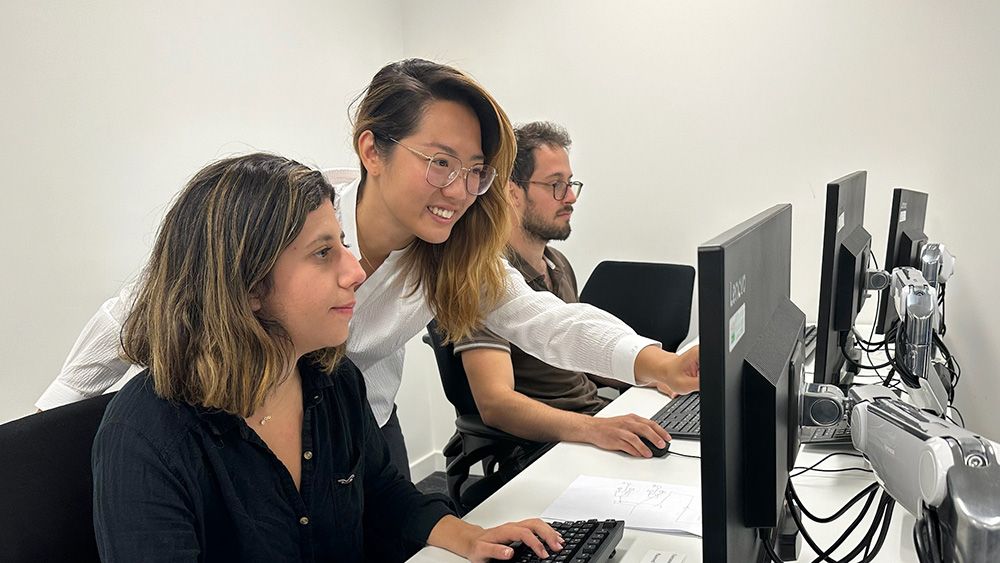 Brenda leans over the shoulder of a woman seated in front of a computer and points at the monitor. In the background a man seated at the same table works on a separate computer.
