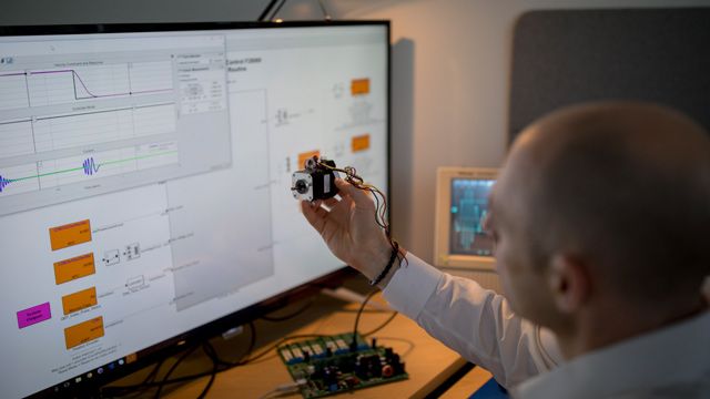 A man holds a piece of hardware while looking at a Simulink model on a screen